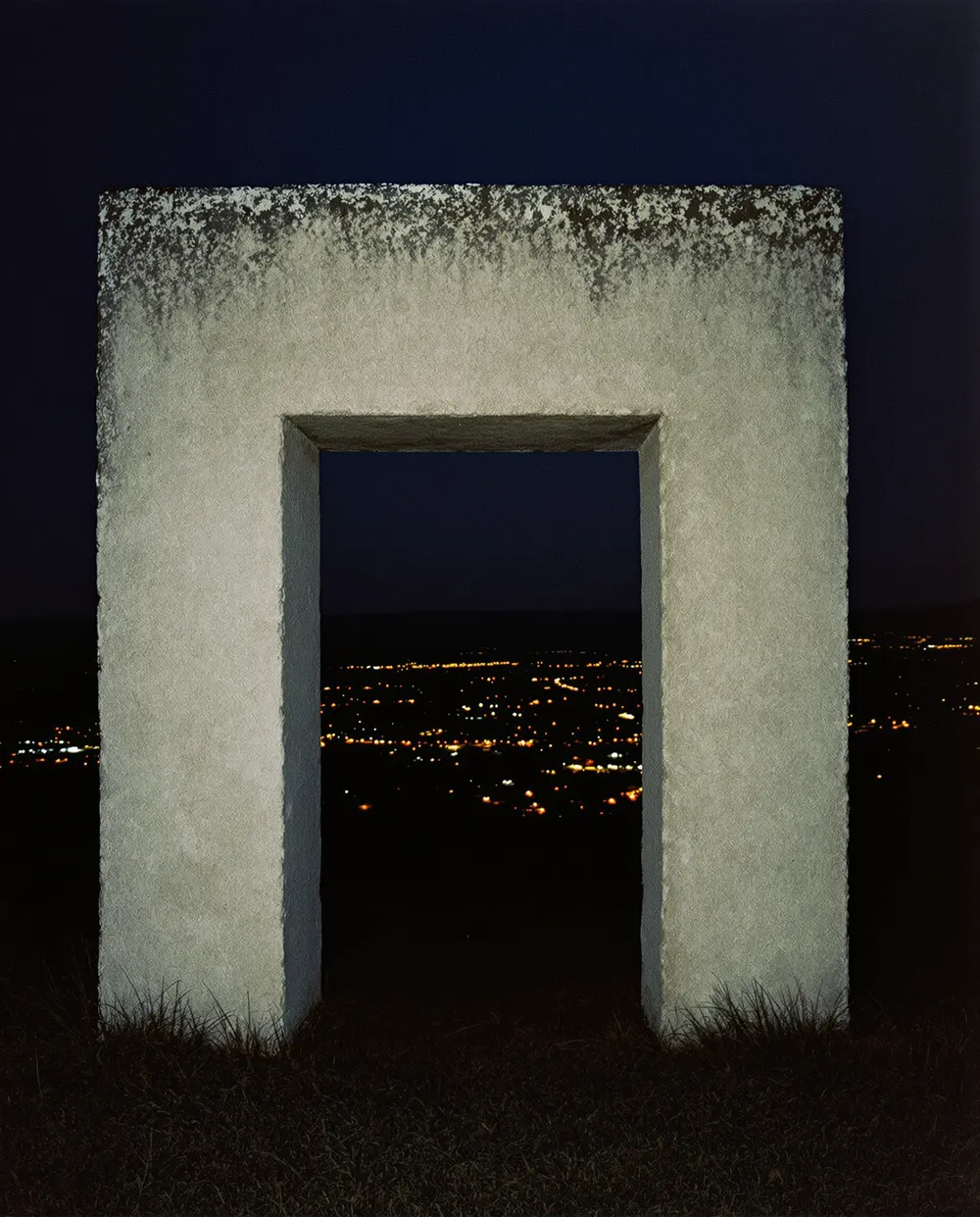 A stone archway standing alone on a hilltop at dusk, the lights of a town visible through the opening.
