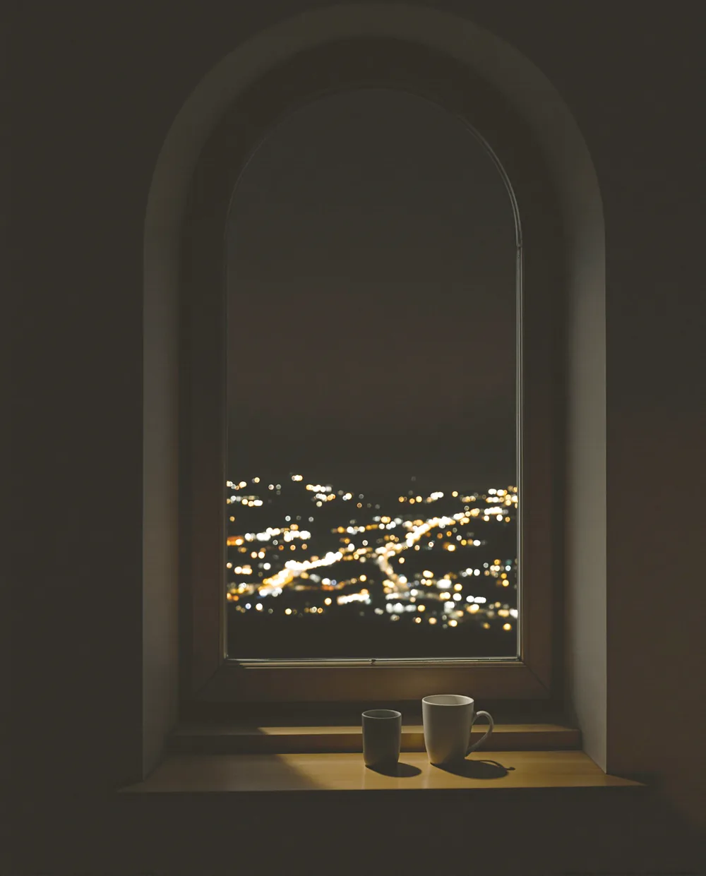 A tall arched window at night seen from inside a dark room, two mugs on the sill, the lights of a town in the valley below.